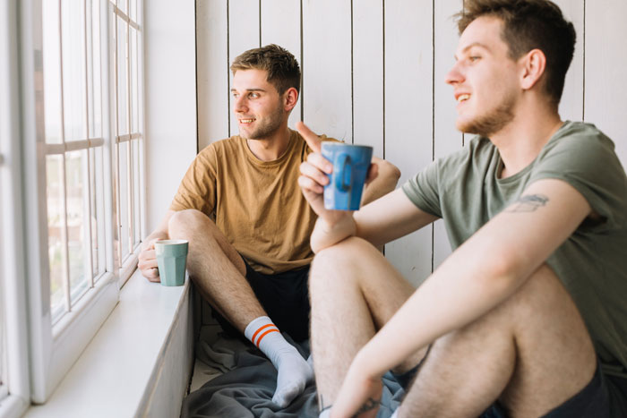 Two young men sitting by a window drinking coffee, illustrating support during a difficult coworker divorce situation.