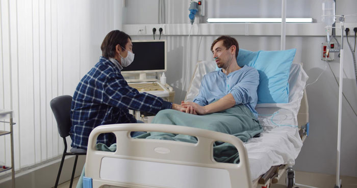 Man recovering from surgery in hospital bed holding hands with masked visitor showing support during difficult time.