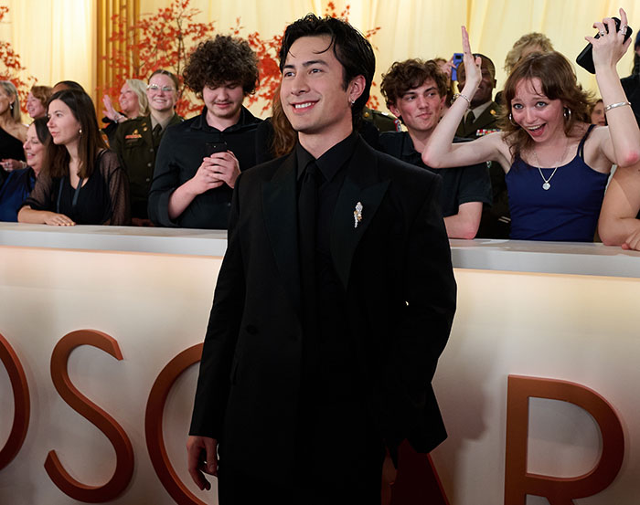 Young man in a black suit smiling at the 2026 Oscars with an excited crowd in the background during cringe moments at the event