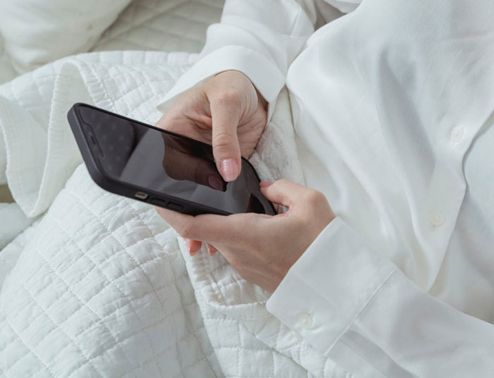 Woman in white shirt holding a smartphone in bed, symbolizing uncovering husband's affair and life turning upside down.