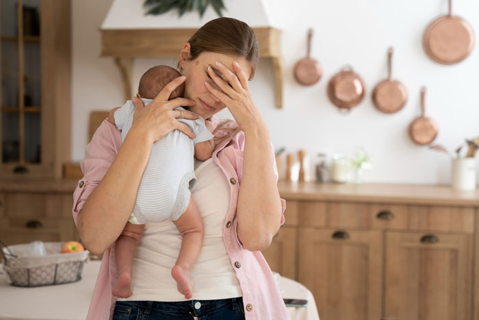 Stressed woman holding baby in kitchen, depicting conflict and sabotage in a troubled marriage situation.