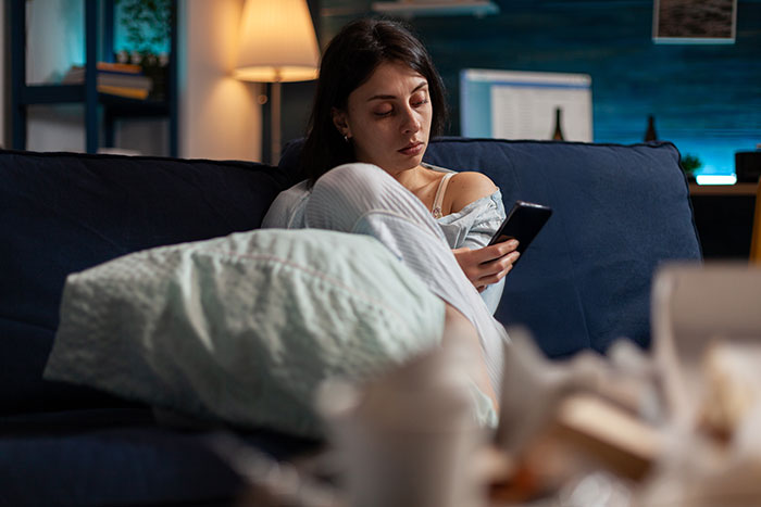 Woman looking concerned while checking phone on couch at night, revealing obsession with sister&rsquo;s weight through texts.
