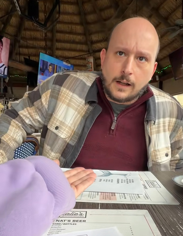 Man in plaid shirt and maroon sweater showing surprise while sitting at a table, illustrating dramatic twist on Tinder.