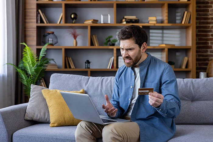 Man sitting on couch holding credit card, looking frustrated at laptop screen about wife sending money to pyramid scheme.