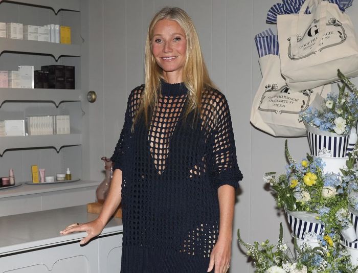 Gwyneth Paltrow smiling at an event, wearing a black mesh top, standing by decorated floral displays and shelves.