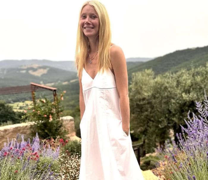 Gwyneth Paltrow smiling outdoors in a white dress with a mountainous landscape and flowers in the background.
