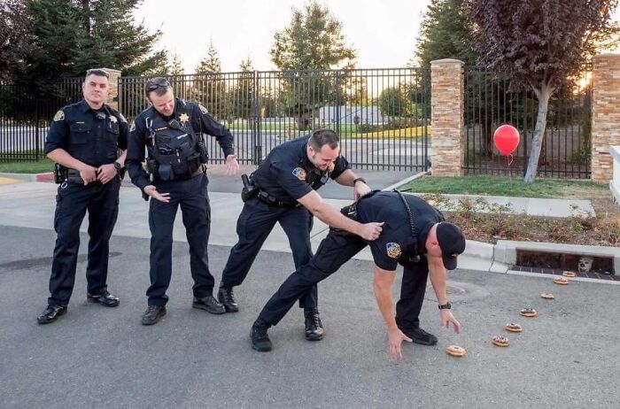 Three police officers laughing while a fourth officer bends over in a hilarious creative pose near donuts on pavement.