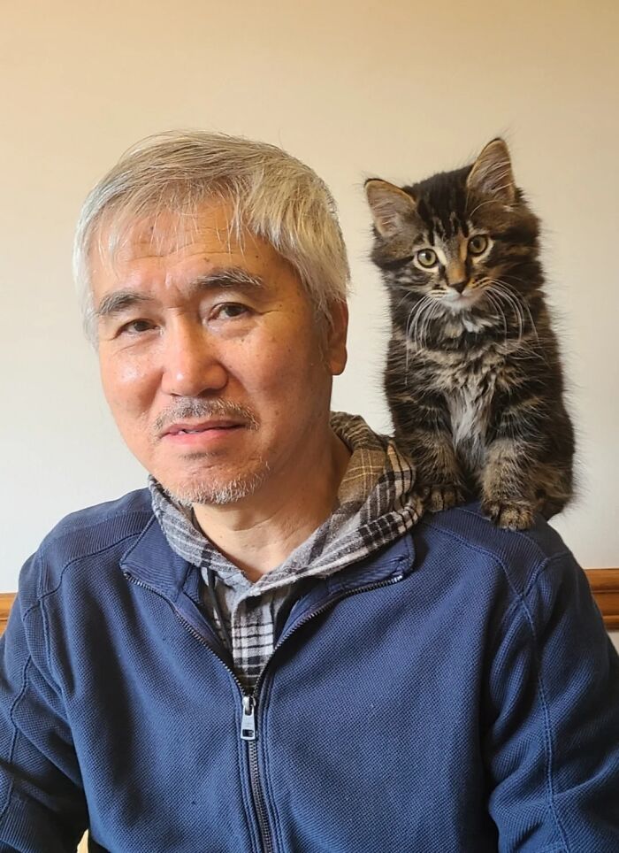 Older man with gray hair wearing a blue jacket with a tabby kitten perched on his shoulder, showing dads and pets love.