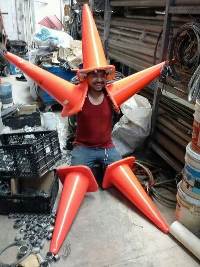 Man sitting on floor wearing multiple orange traffic cones in a creative and hilarious pose in a cluttered workshop space.