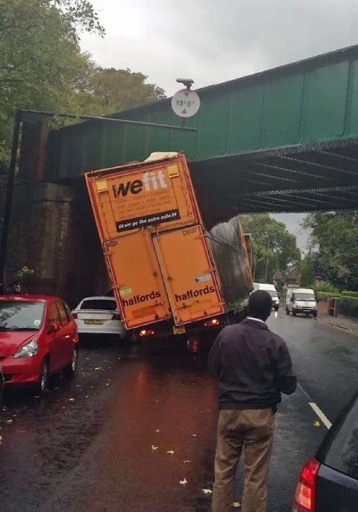 Orange delivery truck stuck under low bridge with man watching on rainy street, illustrating sarcastic images humor in urban setting.