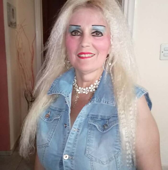 Woman with crazy eyebrows shaped like straight lines, wearing a blue denim vest and long platinum blonde hair indoors.