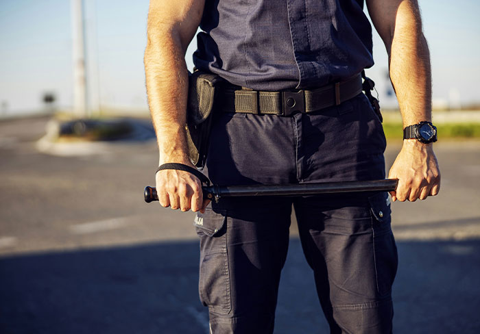 ICE agent in uniform holding a baton standing in a parking lot during a detention incident involving language barriers. ICE agent in uniform holding a baton standing in a parking lot during a detention incident involving language barriers.