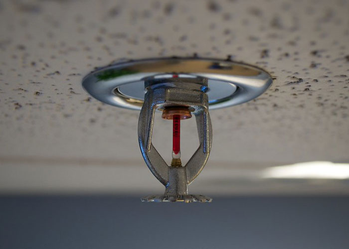 Close-up of a silver fire sprinkler head mounted on a speckled ceiling, illustrating fake facts people believed from movies.