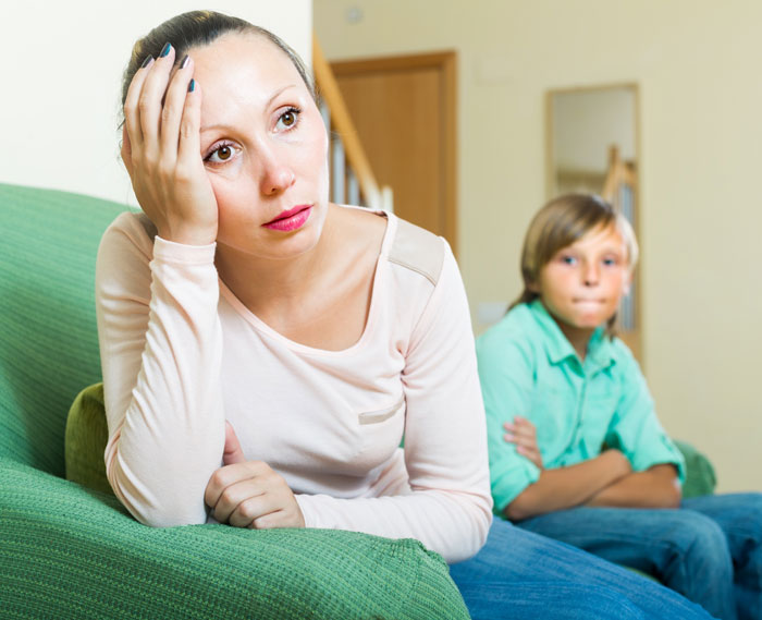 Woman looking distressed on couch while her son sits in the background, highlighting divorced ex and child conflict issues.