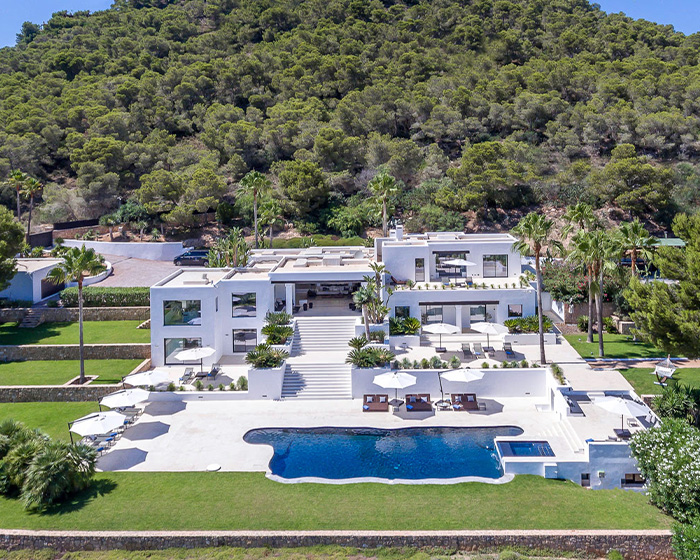 A luxurious modern mansion with a large pool surrounded by palm trees and greenery in a scenic hillside location.