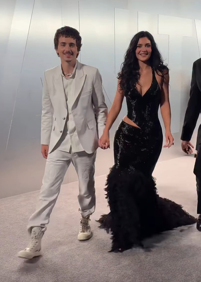 Timoth&eacute;e Chalamet and a woman holding hands, dressed in a white suit and a black gown at an event.