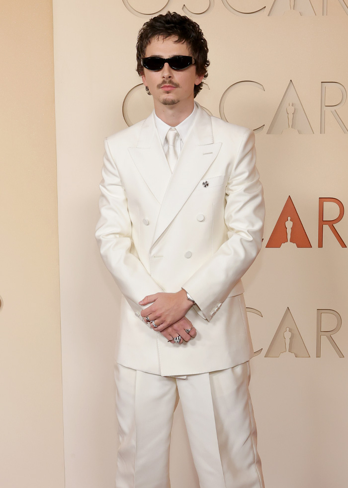 Timoth&eacute;e Chalamet in a white double-breasted suit and sunglasses posing on a beige Oscars event backdrop.