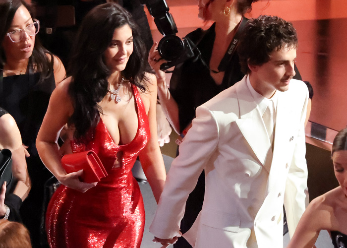 Timoth&eacute;e Chalamet in a white suit holding hands with a woman in a red dress at a crowded event.
