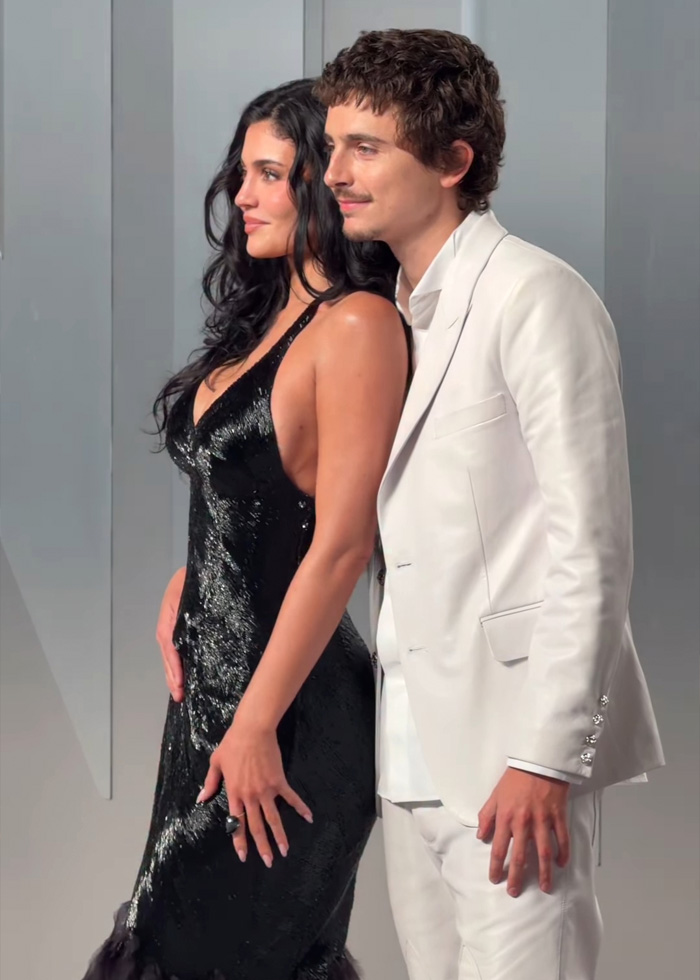 Timoth&eacute;e Chalamet in a white suit posing with a woman in a black dress during an event photoshoot.