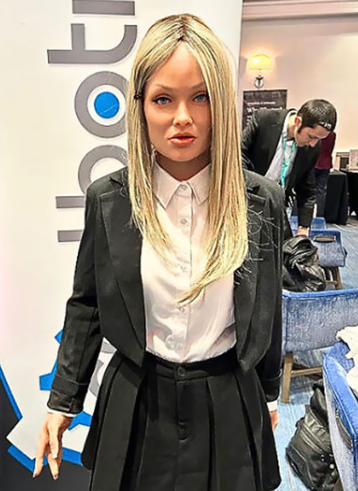 Lifelike robot with blonde hair and business attire at a tech event showcasing strange earth technology innovations.