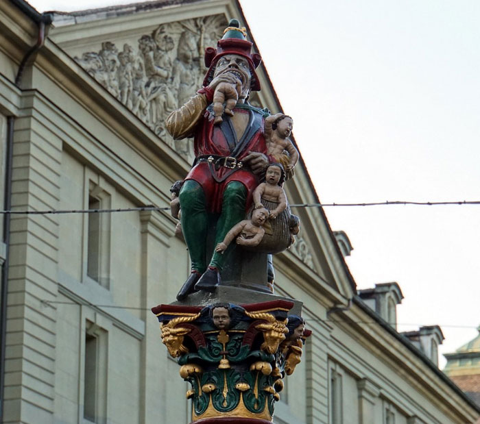 Colorful strange Earth statue of a man holding multiple babies atop an ornate column in an urban setting.