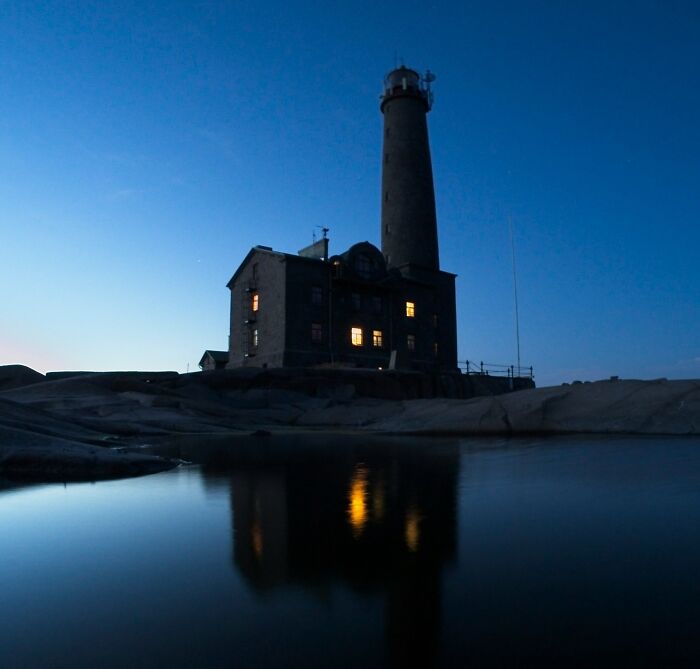 Lighthouse at dusk with lights on, reflecting in water, evoking mystery and unexplained phenomena by the shore.