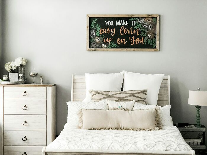 Cozy bedroom with white bedding, pillows, a wooden dresser, and wall art, evoking calm despite haunted places themes.