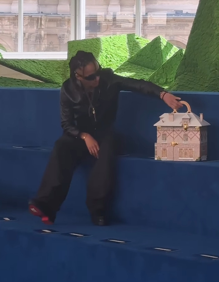 Jaden Smith wearing black outfit and sunglasses sitting on blue steps reaching toward a house-shaped bag.