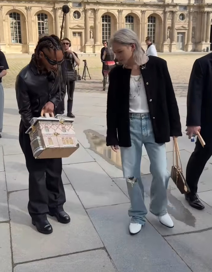 Person in black outfit holding a house-shaped bag while walking outside, with others nearby during a casual day.