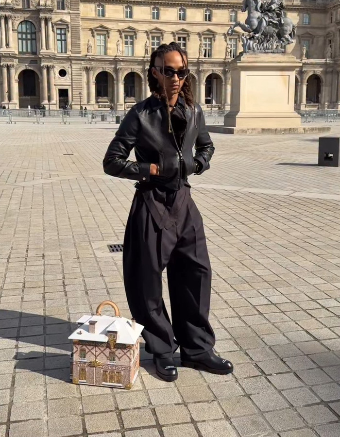 Jaden Smith wearing black leather jacket and sunglasses standing next to a house-shaped bag outside a historic building.