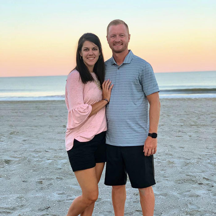 Couple standing on beach at sunset, highlighting heartbreaking details after high school teacher loses life in prank gone wrong.
