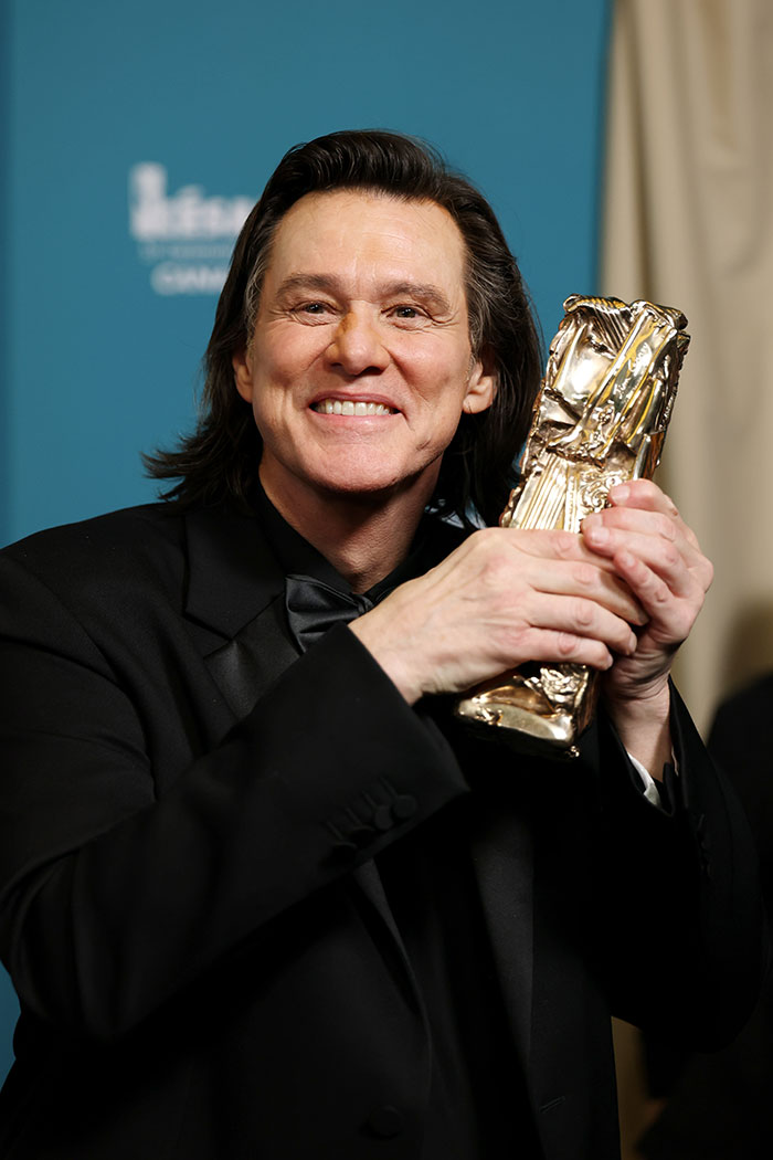 Jim Carrey smiling in a black suit, holding a golden award, highlighting details of his new face and sadness.