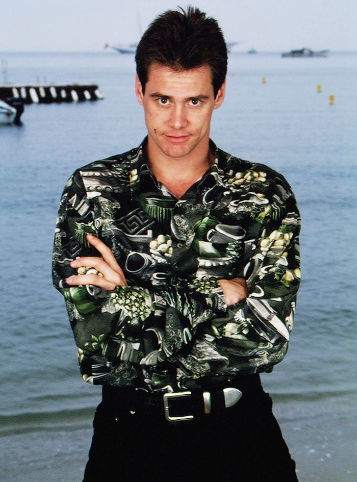 Jim Carrey standing by the water wearing a patterned shirt, arms crossed, with a confident expression and tousled hair.