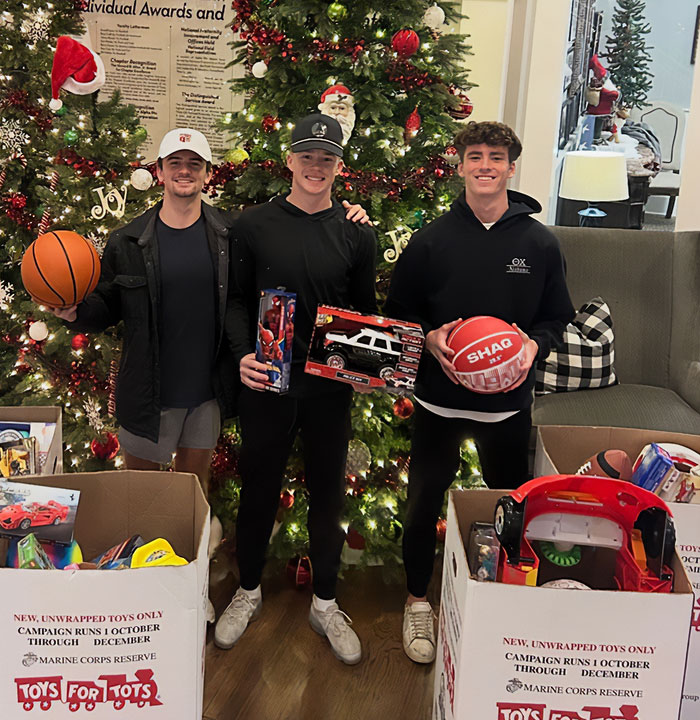 Three young men holding toys and basketballs, standing by decorated Christmas trees and donation boxes.