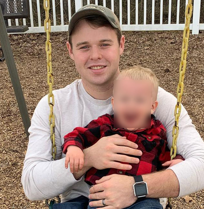 Joseph Duggar sentado em um balanço ao ar livre segurando uma criança vestindo uma camisa xadrez vermelha em um playground.