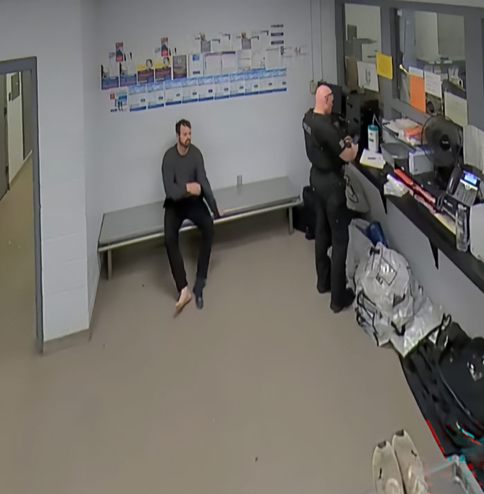 Homem sentado em um banco dentro de uma sala de detenção enquanto um policial fica próximo na filmagem da prisão da esposa de Joseph Duggar, Kendra.