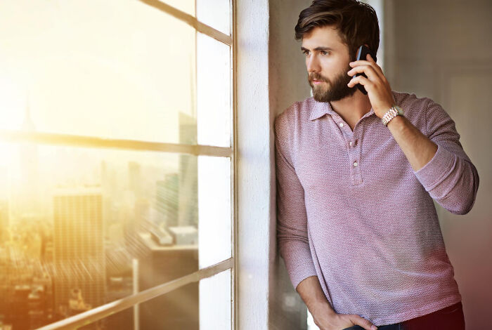 Man with beard leaning against wall by window, talking on phone, reflecting on justice system and jury room experiences.