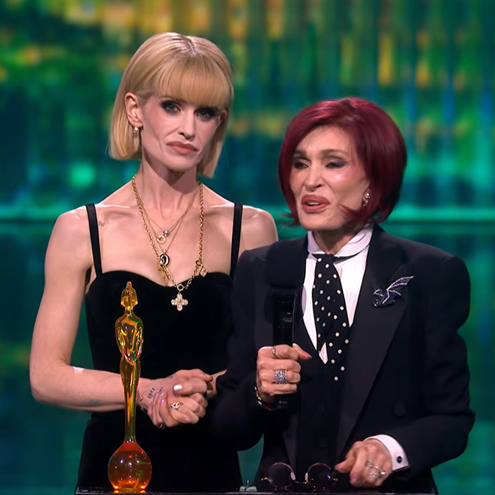 Kelly Osbourne wearing a black dress with layered necklaces standing next to a woman in a black suit at an award event.