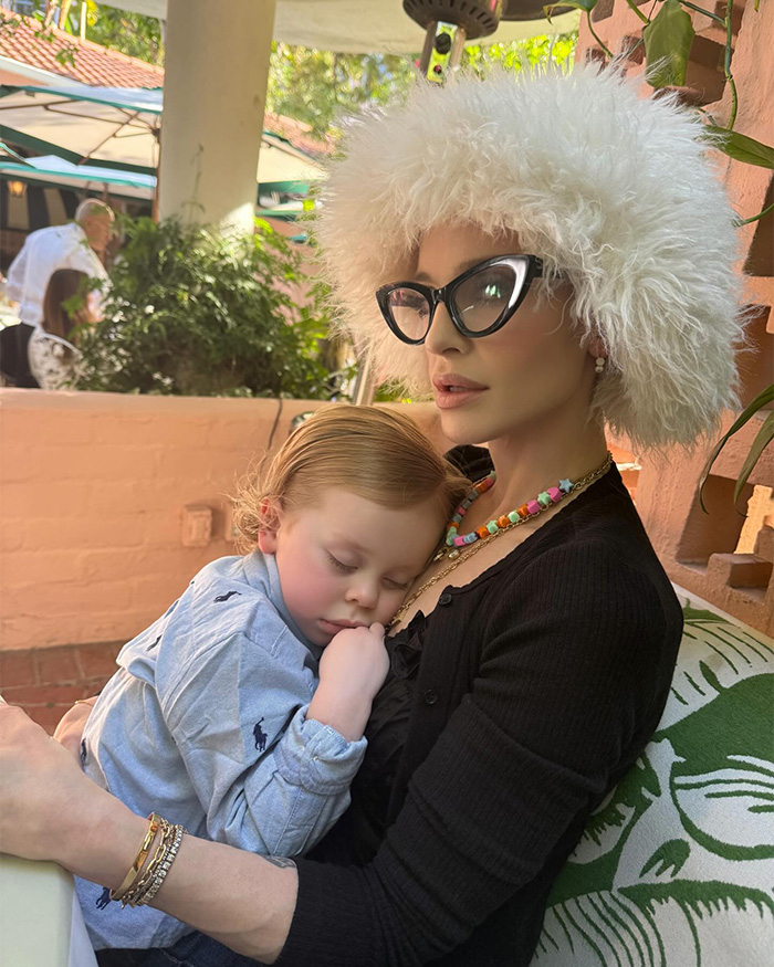 Kelly Osbourne wearing a fluffy white hat and glasses, holding a sleeping child, showcasing her dramatic transformation.