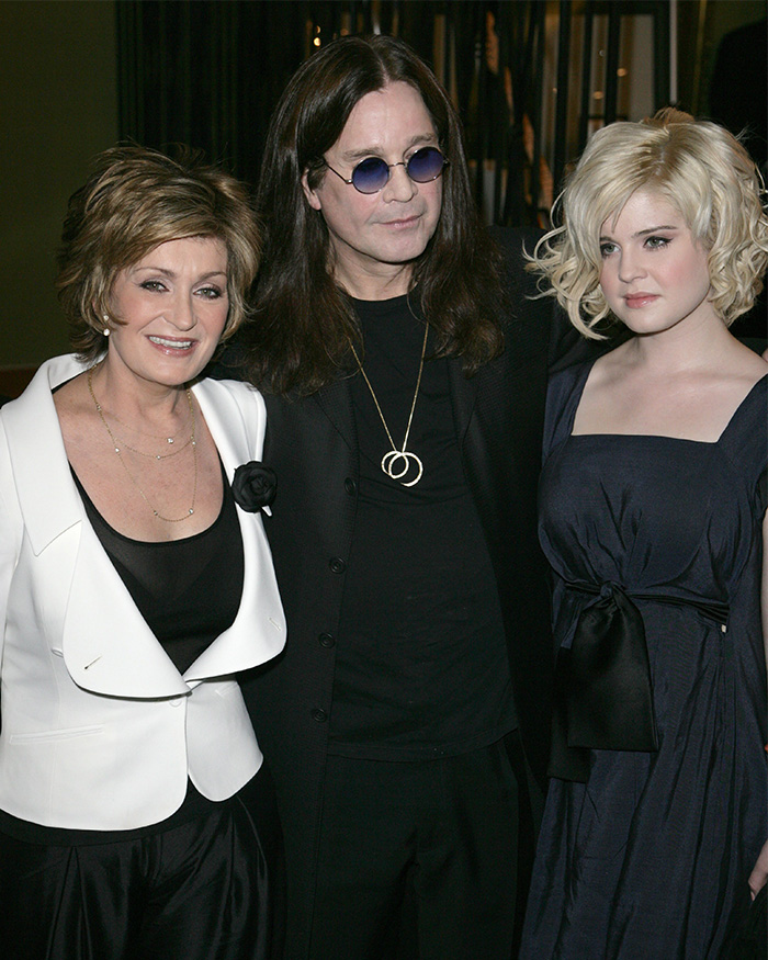 Kelly Osbourne posing with two people at an event, wearing a dark dress, highlighting her dramatic transformation.