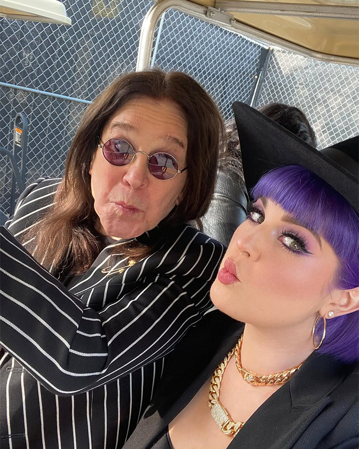 Kelly Osbourne with purple hair and bold makeup posing with a man in sunglasses and a striped jacket outdoors.
