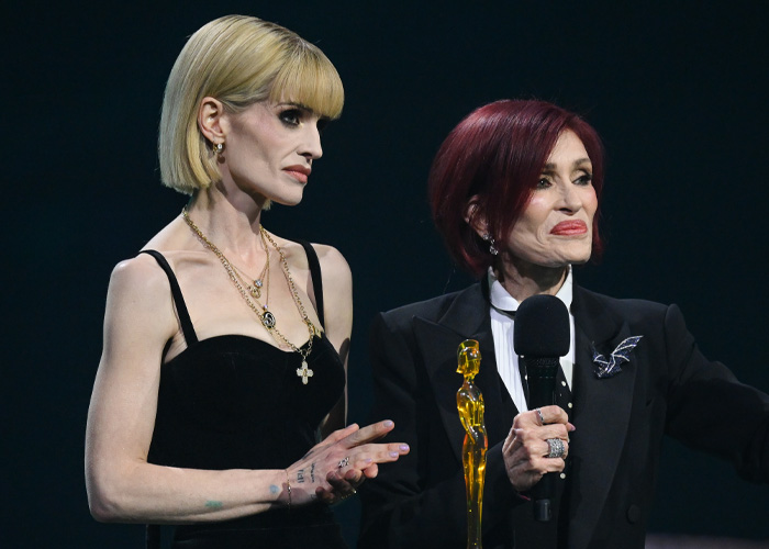 Kelly Osbourne at an event wearing a black dress and gold necklaces, responding to trolls after Brit Awards look goes viral.