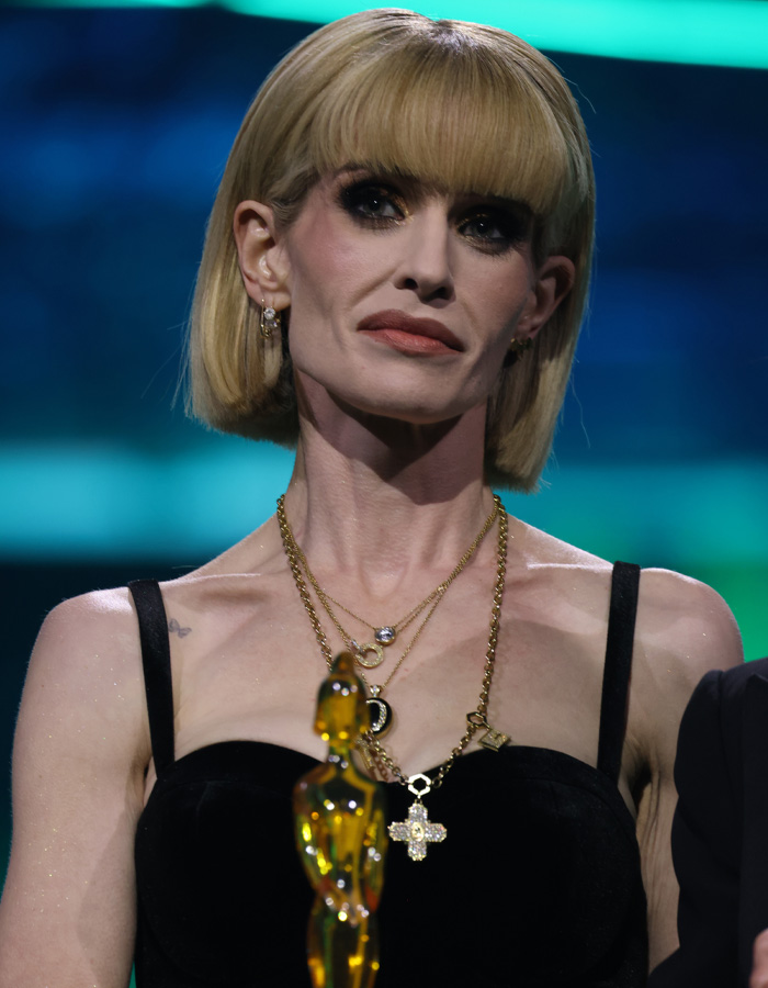Kelly Osbourne wearing layered gold necklaces and black dress reacting at an event amid Brit Awards trolls controversy.