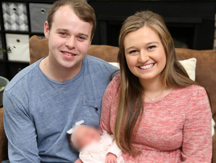A esposa de Joseph Duggar, Kendra, sorrindo com o marido e o bebê em um ambiente doméstico aconchegante, relacionada a notícias de imagens de prisão.