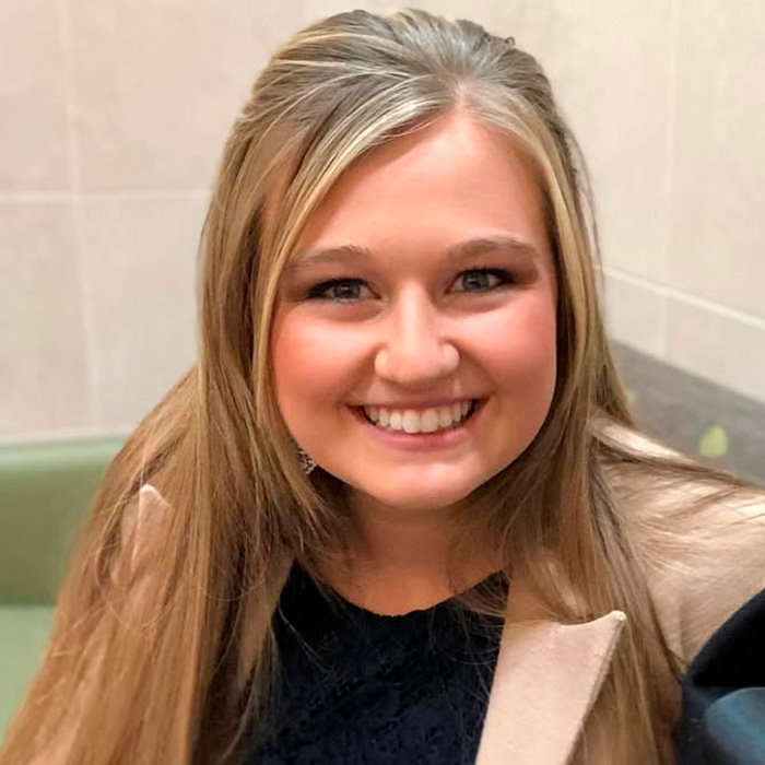 Kendra Duggar smiling with long hair wearing a beige coat and black top in a casual indoor setting.