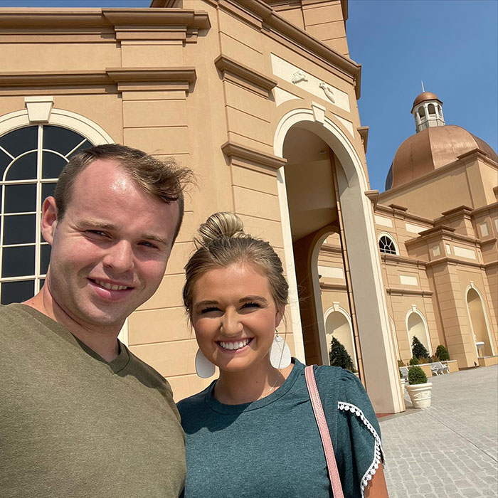 Joseph Garrett Duggar with a woman, smiling outdoors near a large beige building under clear blue sky.