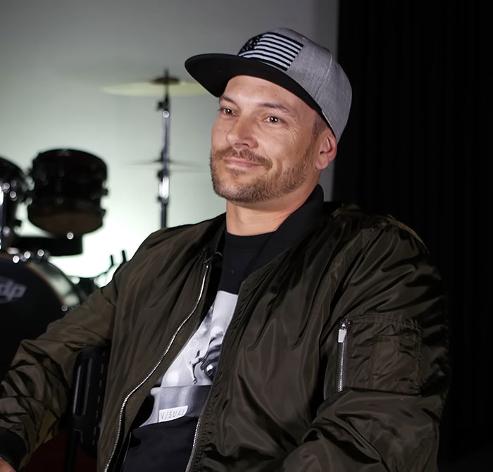 Man wearing a cap and jacket sitting calmly with drums in the background, related to Britney Spears arrest report.