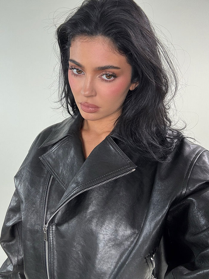 Kylie Jenner posing in a black leather jacket, showing natural makeup and a serious expression in a studio setting.