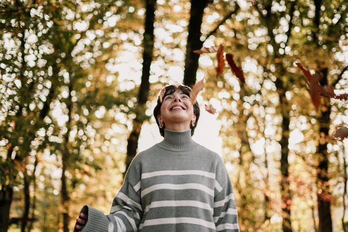 Person enjoying autumn in a forest, smiling and surrounded by falling leaves in a calm outdoor setting, lifestyle concept.