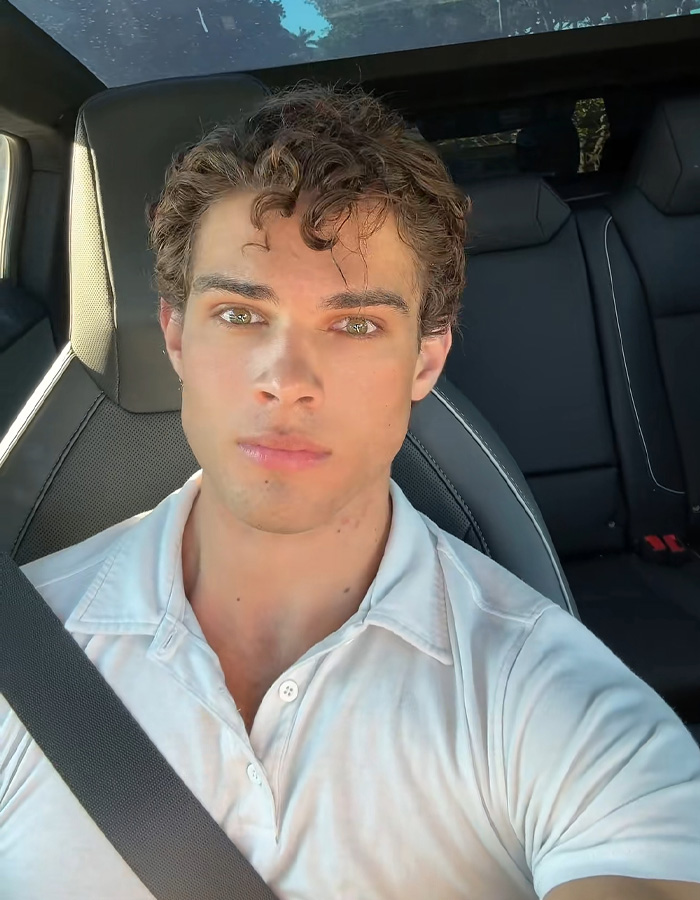 Young man with curly hair and green eyes wearing a white shirt, sitting in a car seat with a seatbelt, looksmaxxing influencer.